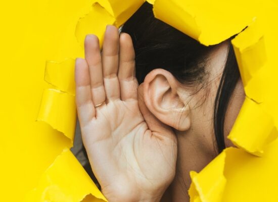 Close-up of a woman's ear and hand through a torn hole in the paper. Bright yellow background, copy space. The concept of eavesdropping, espionage, gossip and tabloids.