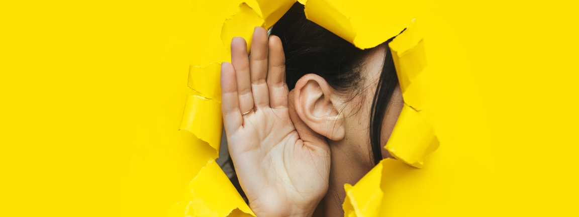 Close-up of a woman's ear and hand through a torn hole in the paper. Bright yellow background, copy space. The concept of eavesdropping, espionage, gossip and tabloids.