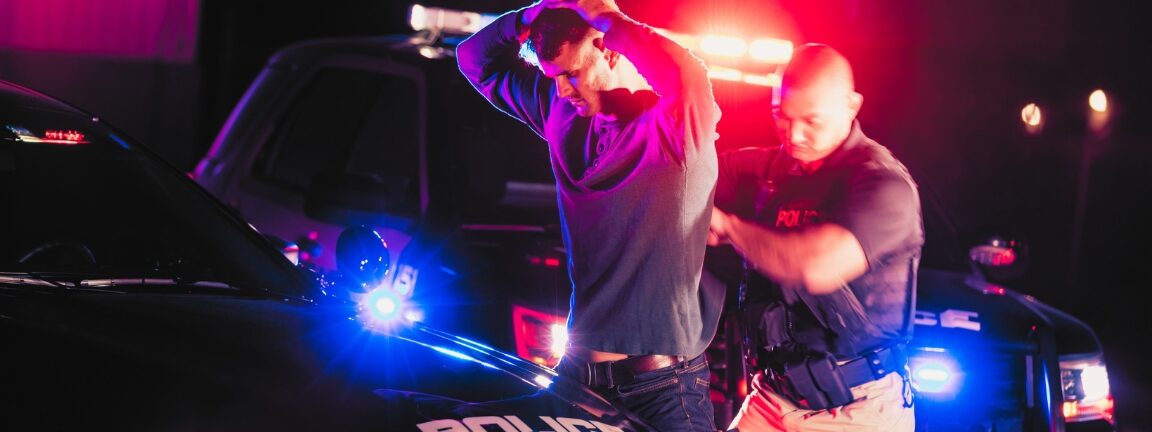 A police officer making an arrest, standing outside his patrol car at night, emergency lights flashing. The criminal is a young man in his 20s, standing his his hands on his head as the officer searches him.