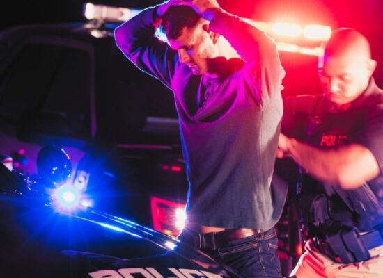 A police officer making an arrest, standing outside his patrol car at night, emergency lights flashing. The criminal is a young man in his 20s, standing his his hands on his head as the officer searches him.